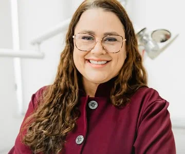 Dra. Fabíola Cassiano de jaleco bordô, sorrindo no consultório.