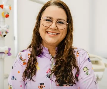 Dra. Fabíola Cassiano de jaleco infantil estampado, sorrindo no consultório com brinquedos ao fundo.
